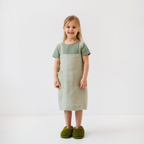 3. Girl smiling in sage green linen pinafore apron with hands in pockets, standing on white background