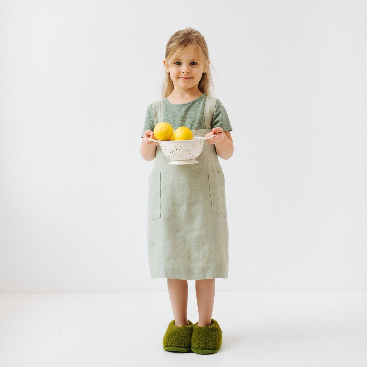 2. Girl in sage green linen pinafore apron holding a colander with lemons, standing on white background