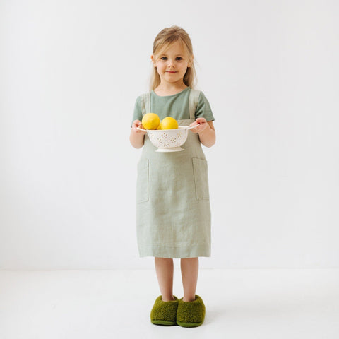2. Girl in sage green linen pinafore apron holding a colander with lemons, standing on white background