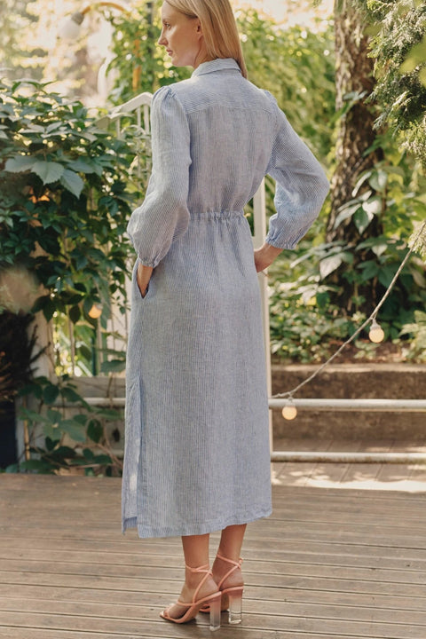 2. Back view of blue and white striped linen dress with side slits and elasticated cuffs, worn by woman on wooden deck