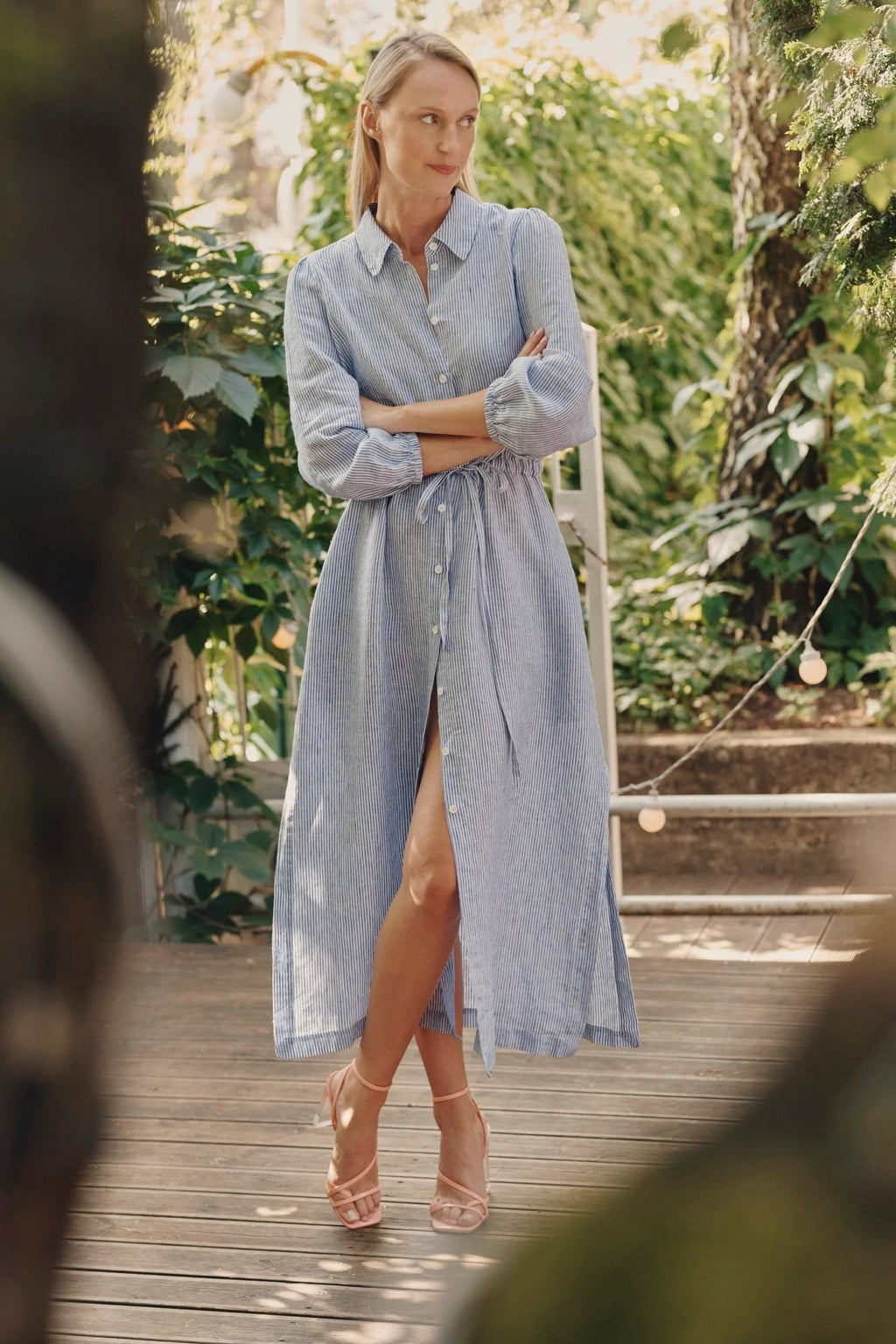 1. Woman wearing blue and white striped linen dress with side slits and adjustable belt, standing on a wooden deck surrounded by greenery