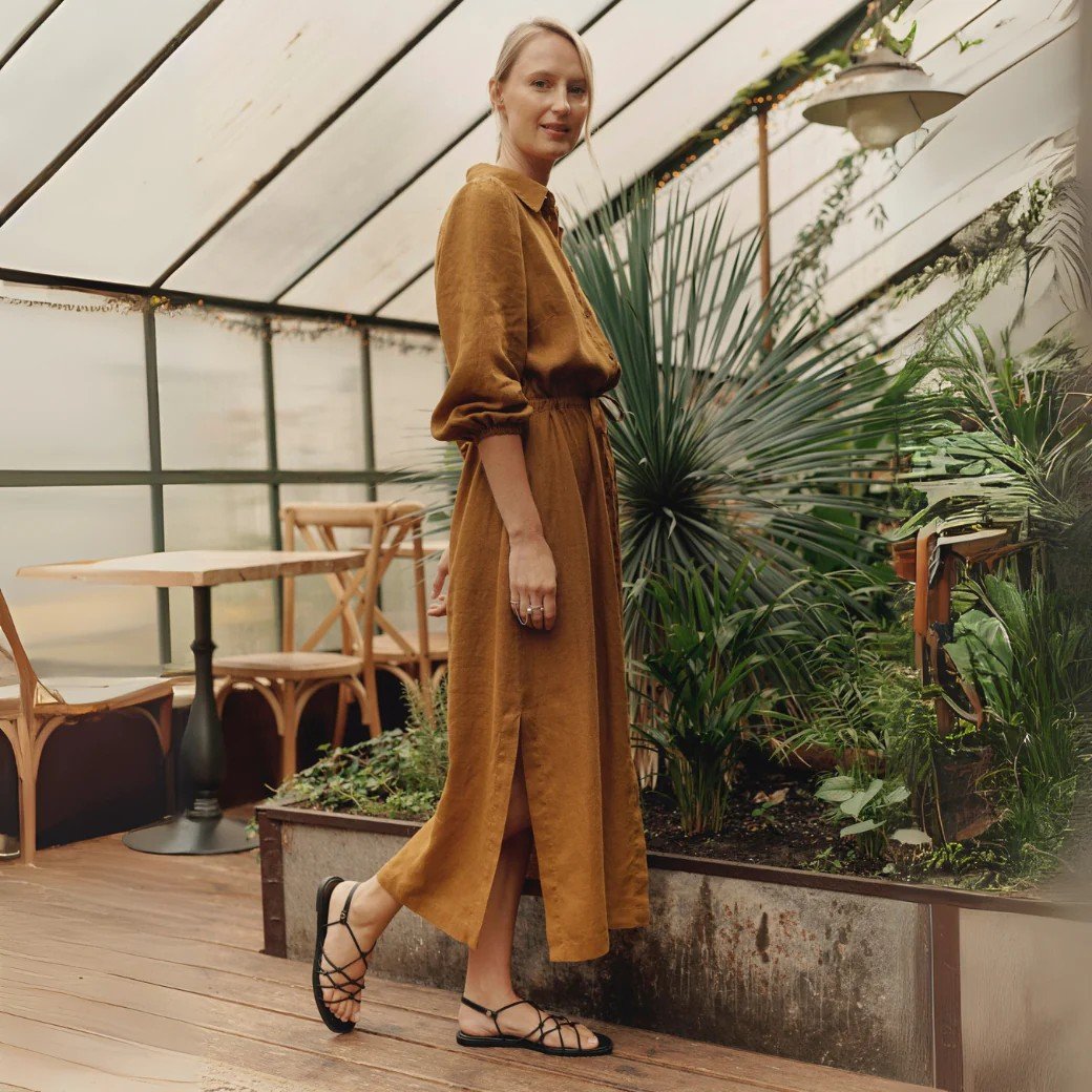 2. Woman in bronze linen Rhapsody dress with side slits and elasticated cuffs, posing in a greenhouse