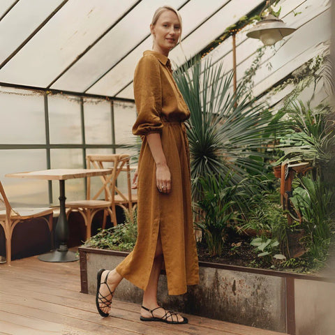 2. Woman in bronze linen Rhapsody dress with side slits and elasticated cuffs, posing in a greenhouse