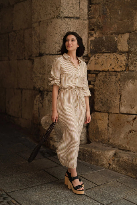 1. Woman wearing bronze and white striped linen dress with side slits and adjustable belt, walking along a stone wall