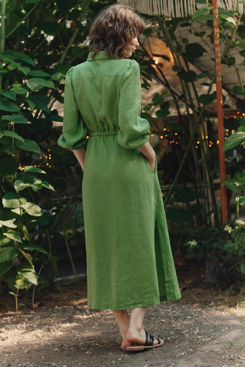 2. Back view of green linen long shirt dress with elasticated cuffs and side slits, worn by woman in garden