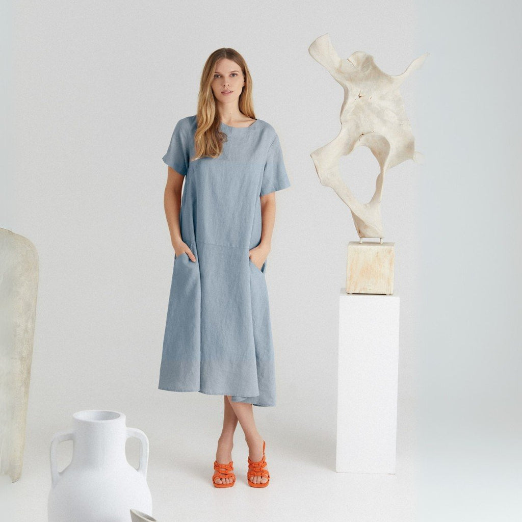 1. Woman wearing blue linen A-line dress with short sleeves and round neck, standing in a minimalist studio setting with abstract sculptures, showcasing front pockets and relaxed fit