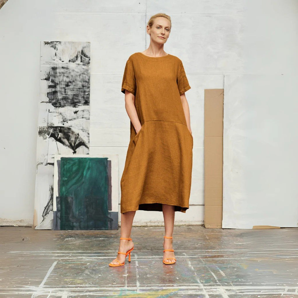 1. Woman wearing bronze linen A-line dress with short sleeves and round neck, standing in an art studio setting with abstract paintings in the background