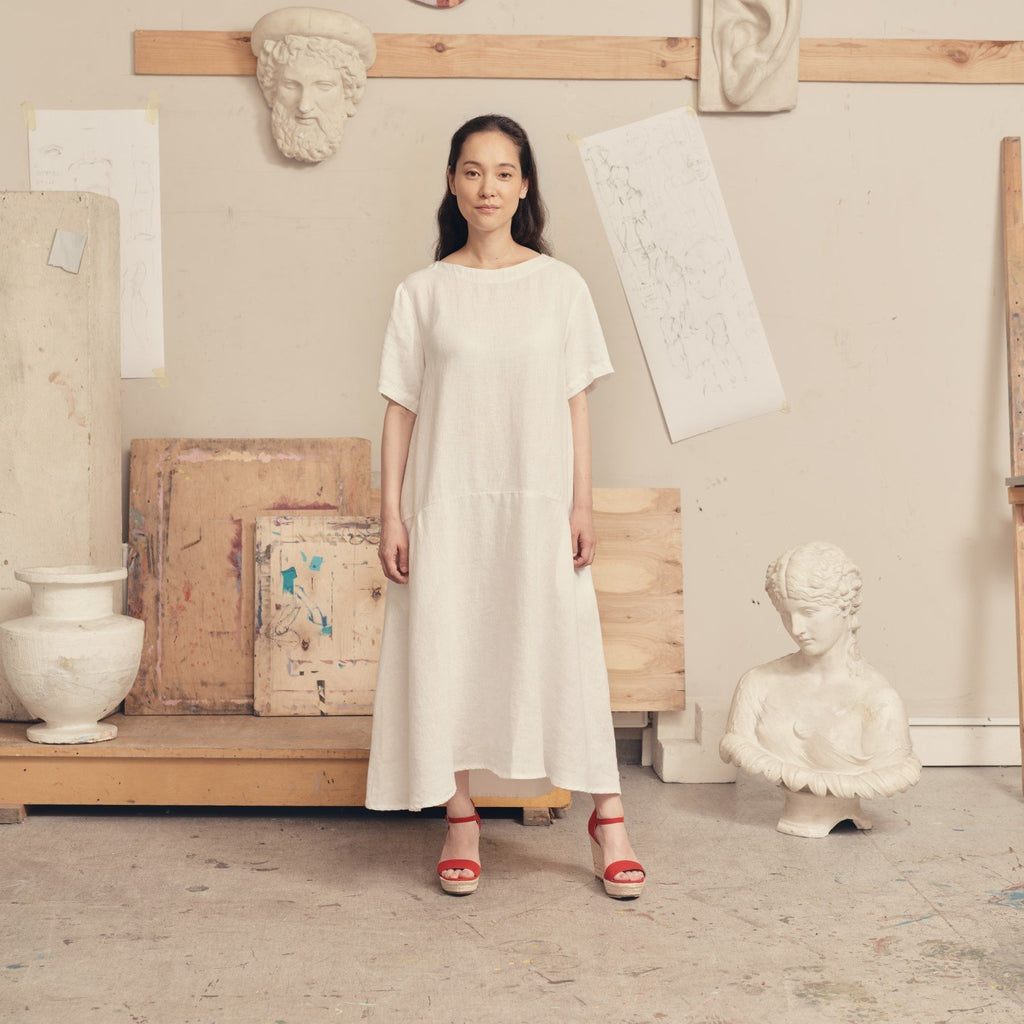 1. Woman wearing optical white linen dress with red sandals in an art studio setting, surrounded by sculptures and sketches
