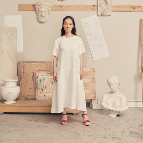 1. Woman wearing optical white linen dress with red sandals in an art studio setting, surrounded by sculptures and sketches