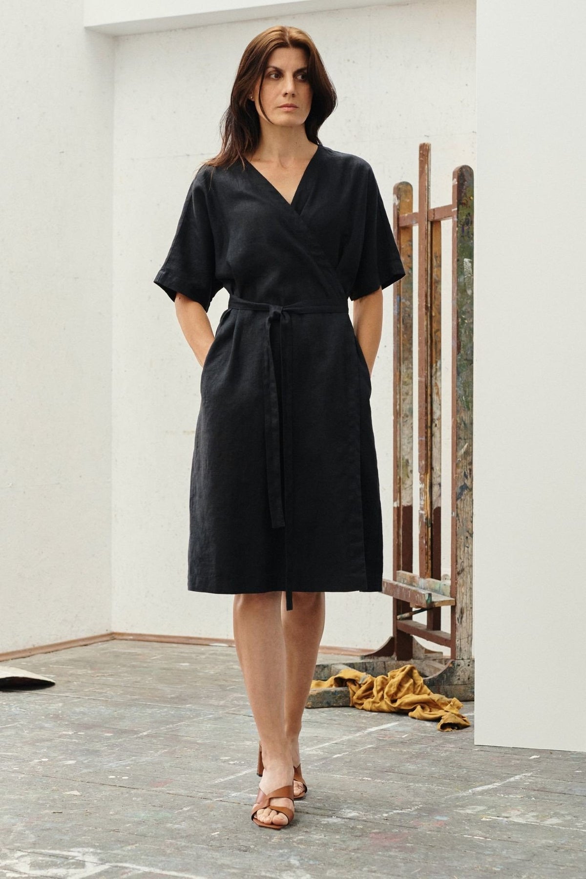 1. Woman wearing black linen wrap dress with V-neck and tied fastening, standing in an art studio setting