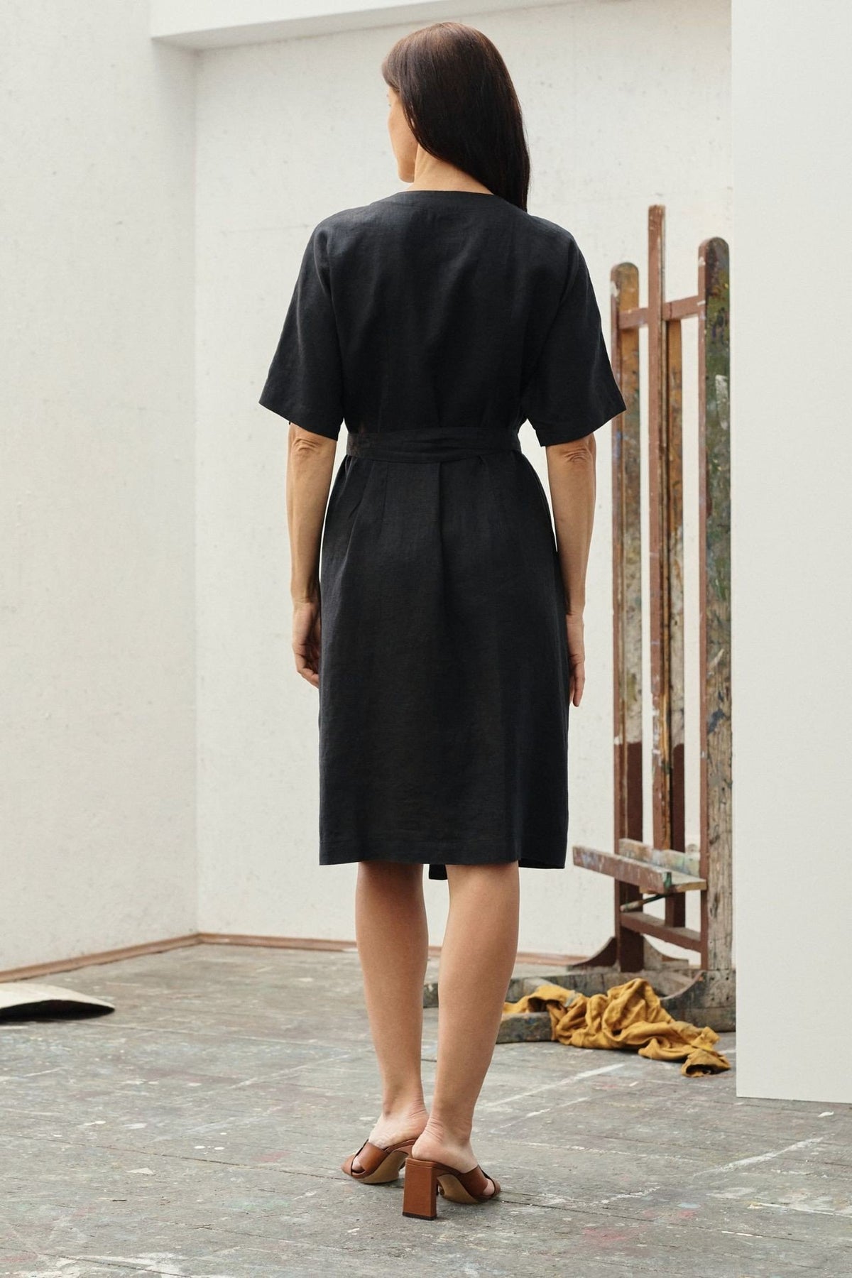 2. Back view of black linen wrap dress with short sleeves and tied waist, worn by a woman in an art studio