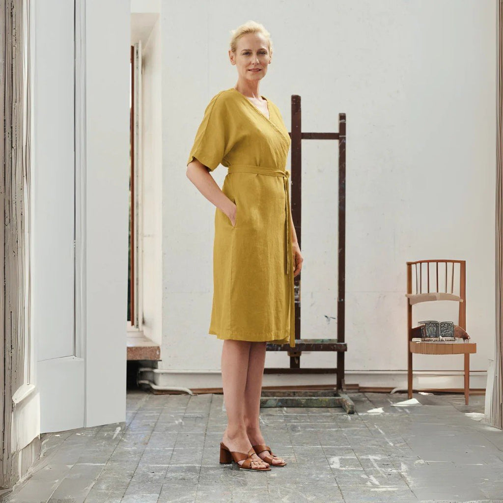 1. Woman wearing lemon curry linen wrap dress with V-neck and tied fastening, standing in a bright room with wooden floors and a chair in the background