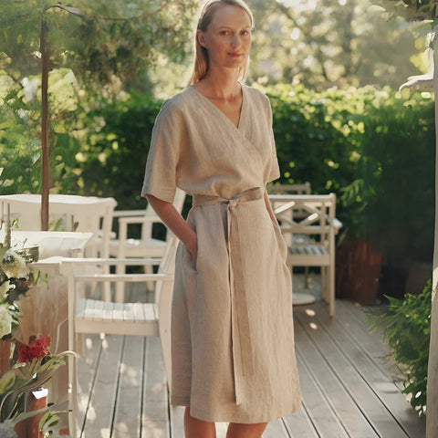 1. Woman wearing beige linen wrap dress with V-neck and tied fastening, standing on a wooden deck surrounded by greenery