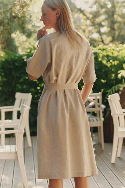 2. Back view of woman in beige linen wrap dress with tied waist, standing on a wooden deck with chairs and greenery