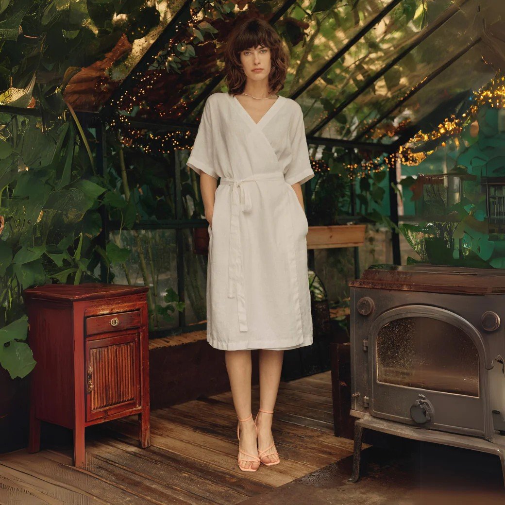 1. Woman wearing optical white linen wrap dress with V-neck and tied fastening, standing in a greenhouse setting with plants and vintage furniture