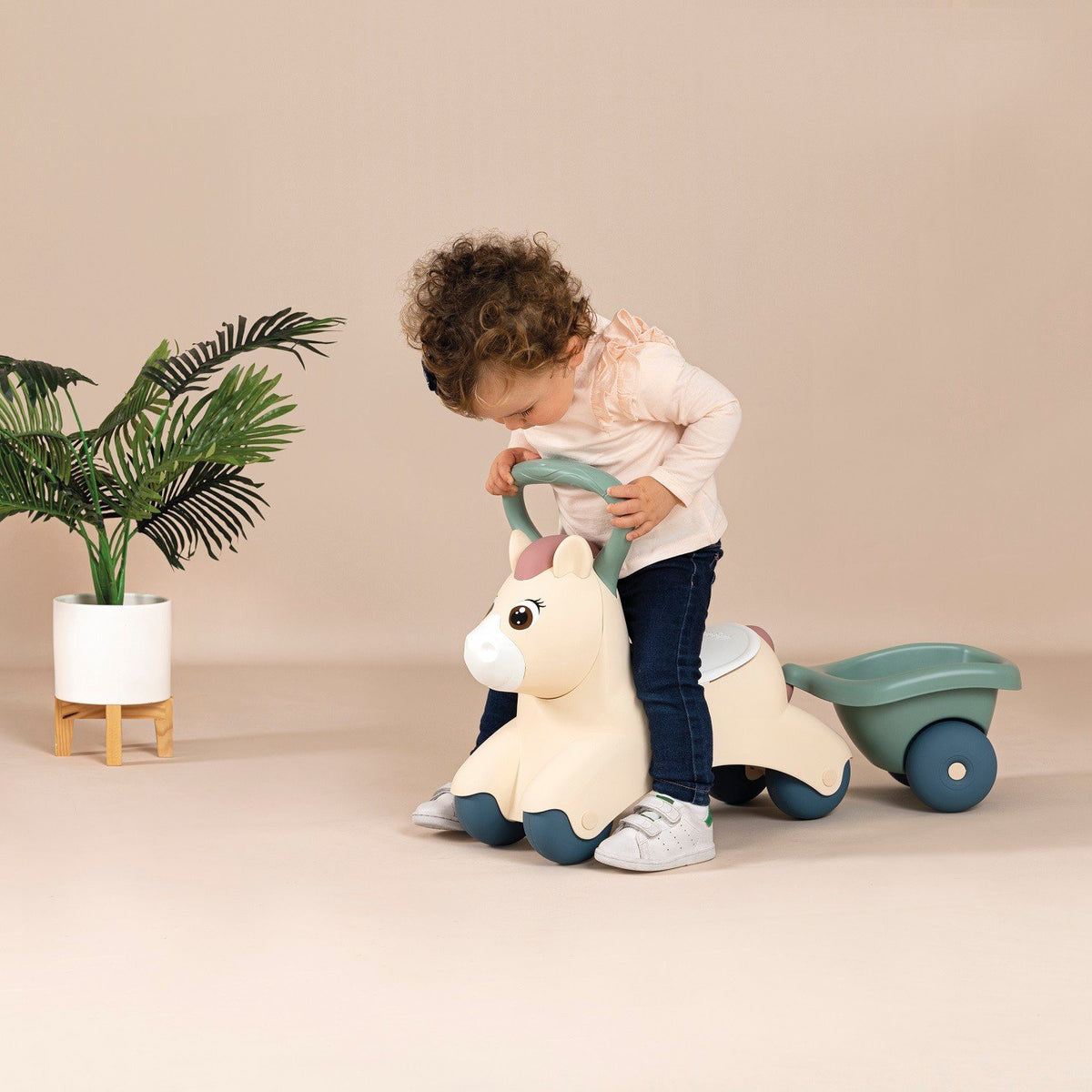 2. Child riding beige pony ride-on with green trailer in a room with a plant