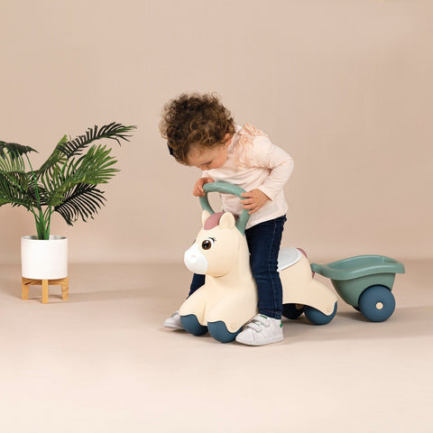 2. Child riding beige pony ride-on with green trailer in a room with a plant