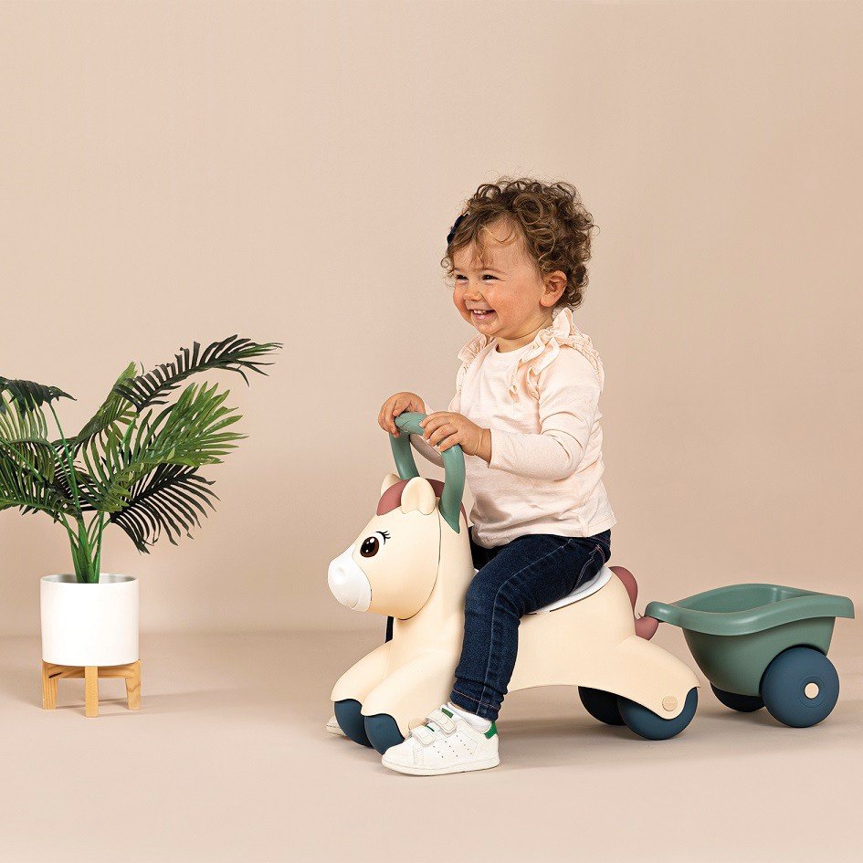 7. Child happily riding beige pony ride-on with green trailer in a room with a plant