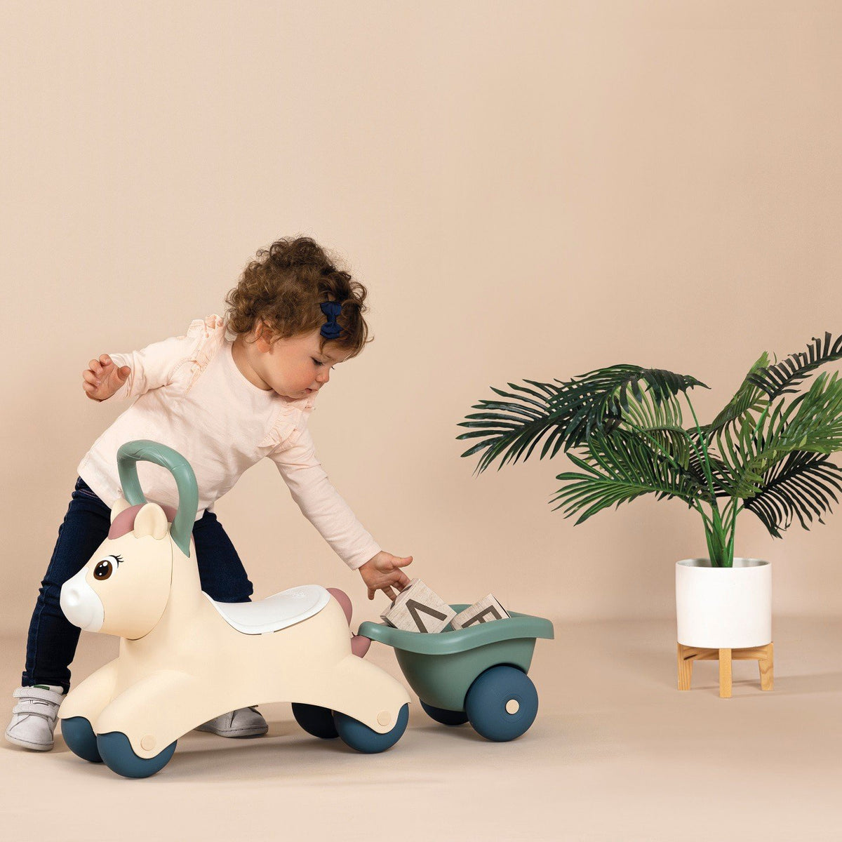 6. Child placing items in green trailer of beige pony ride-on next to a plant