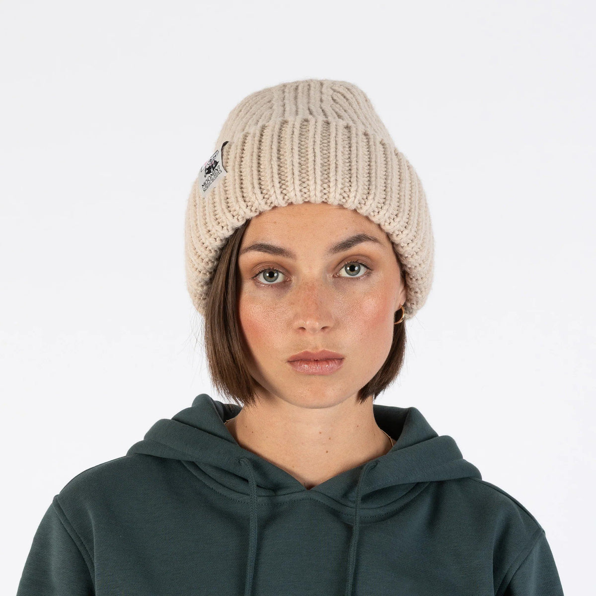 1. Woman wearing beige ribbed beanie with Moomin logo, facing forward, against white background