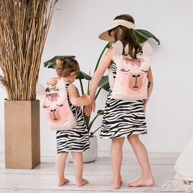 2. Two girls wearing Muni llama backpacks indoors, highlighting size options and cute llama design