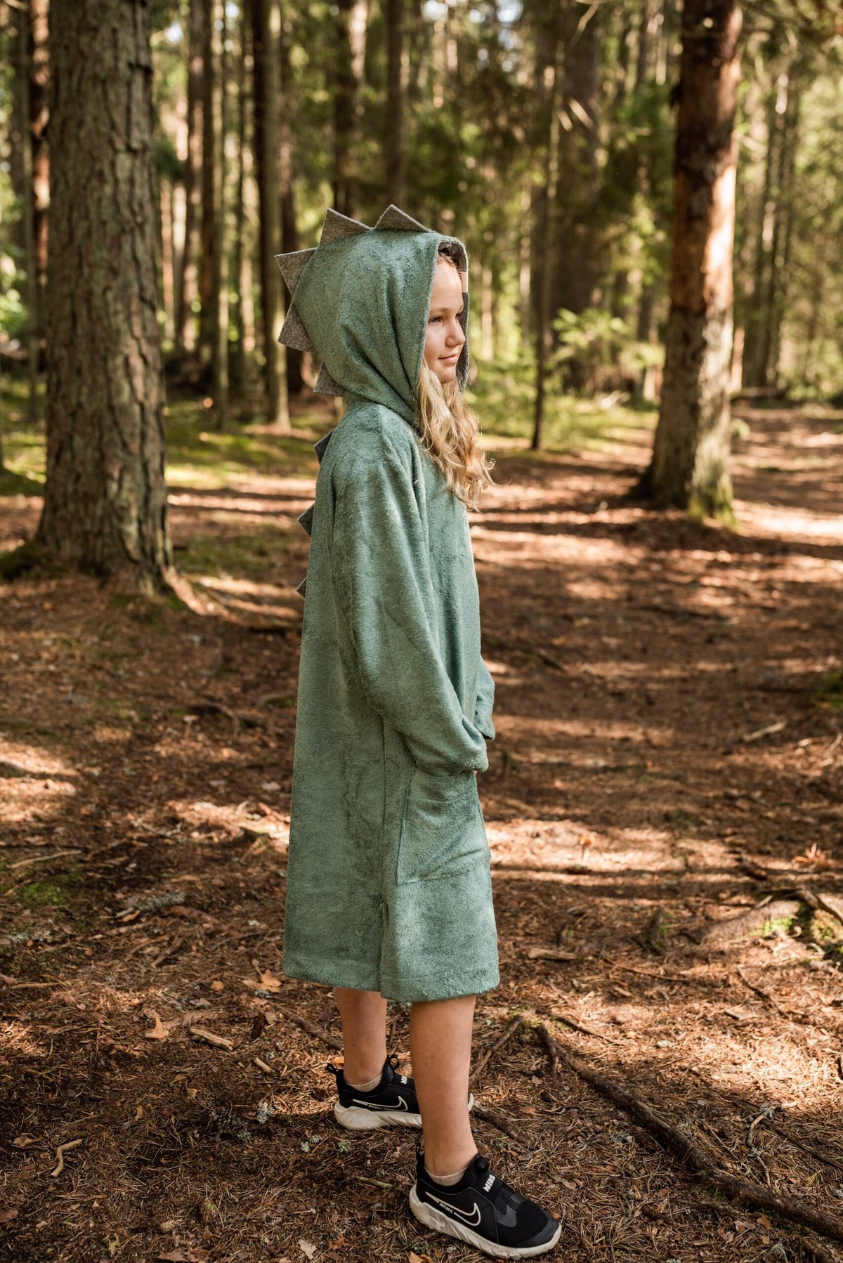 2. Side view of child in RÄTT green dinosaur poncho with hood, standing in a wooded area