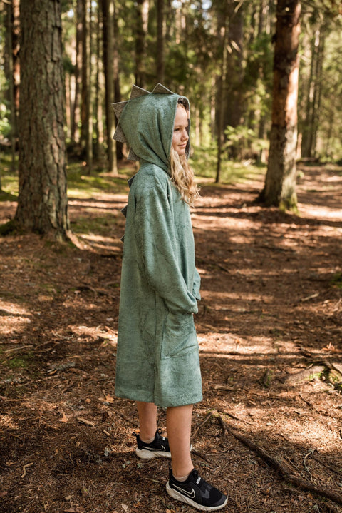 2. Side view of child in RÄTT green dinosaur poncho with hood, standing in a wooded area