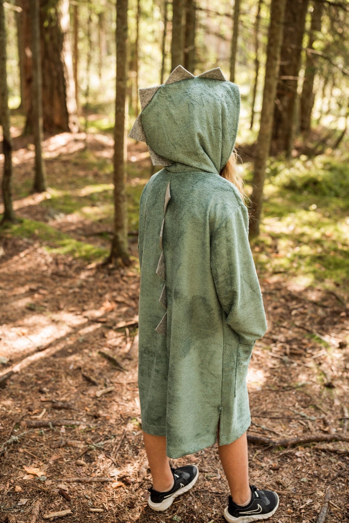 1. Child wearing RÄTT long-sleeve dinosaur poncho in green, shown from the back in a forest setting