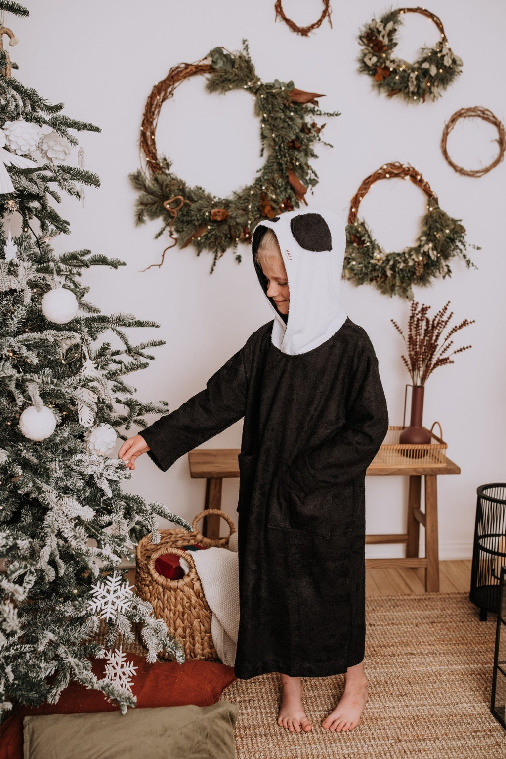 1. Child wearing RÄTT Long-Sleeve Poncho PANDA in black with panda hood, standing by a Christmas tree in a cozy room