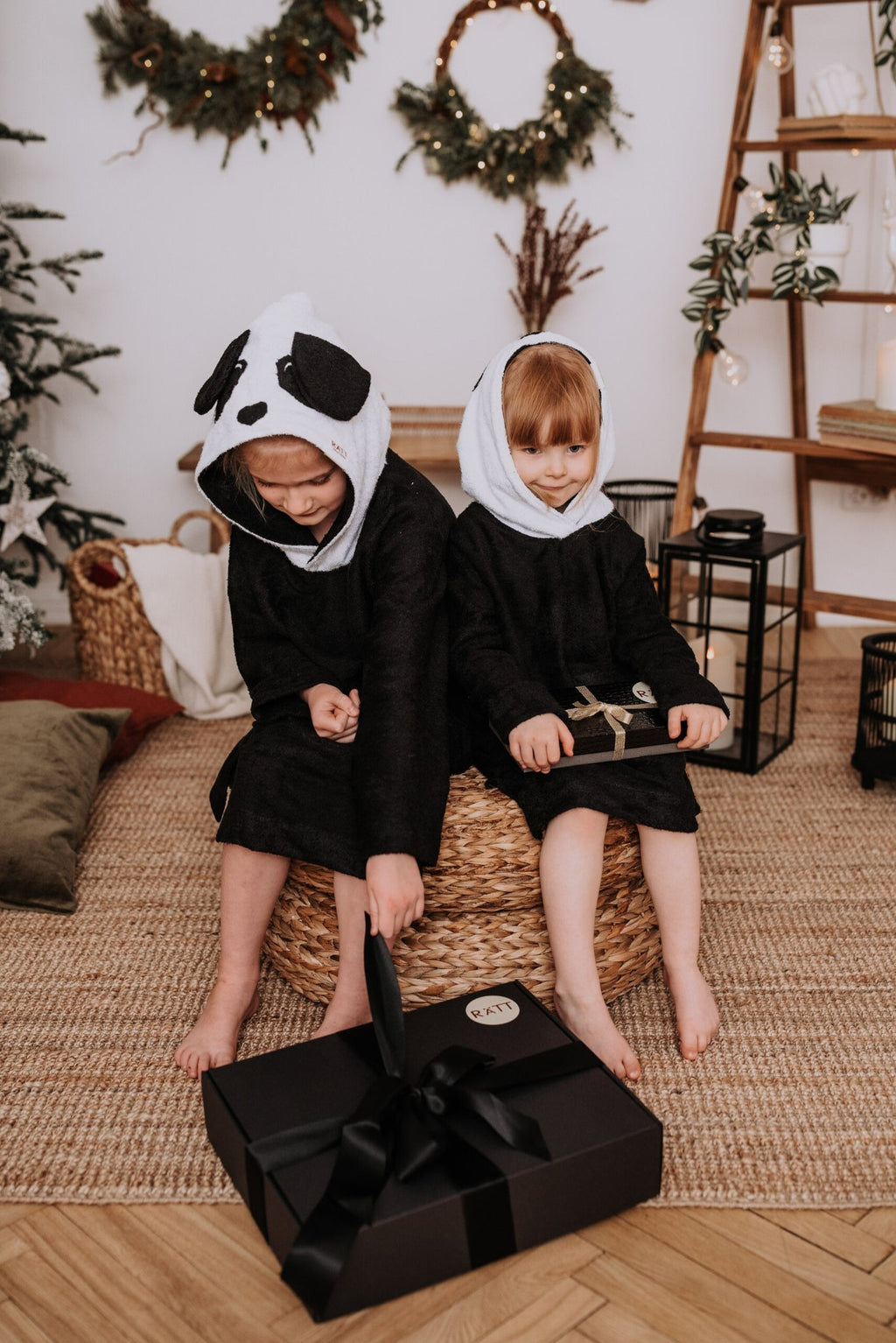 2. Two children sitting together wearing RÄTT Long-Sleeve Poncho PANDA in black with panda hoods, in a festive setting