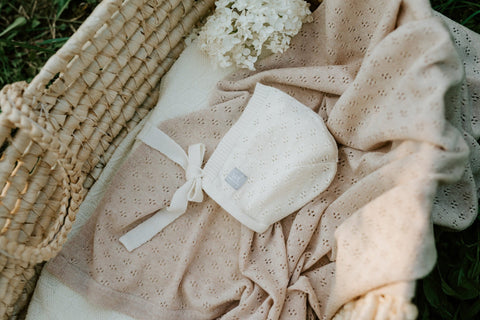 1. Beige merino wool baby blanket and bonnet with floral pattern in a woven basket with flowers