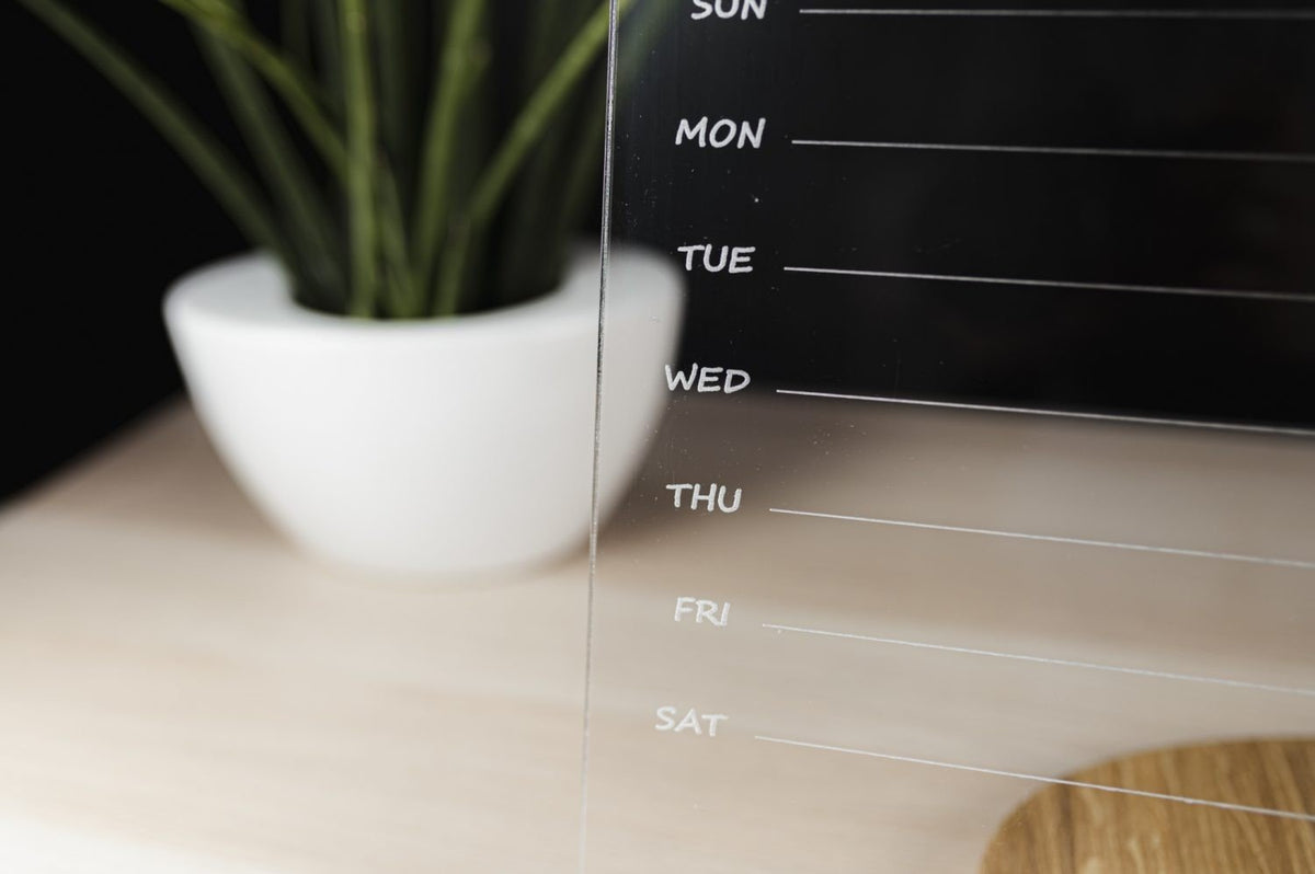 4. Side view of clear acrylic planner showing weekly days and plant in background, on light wood surface