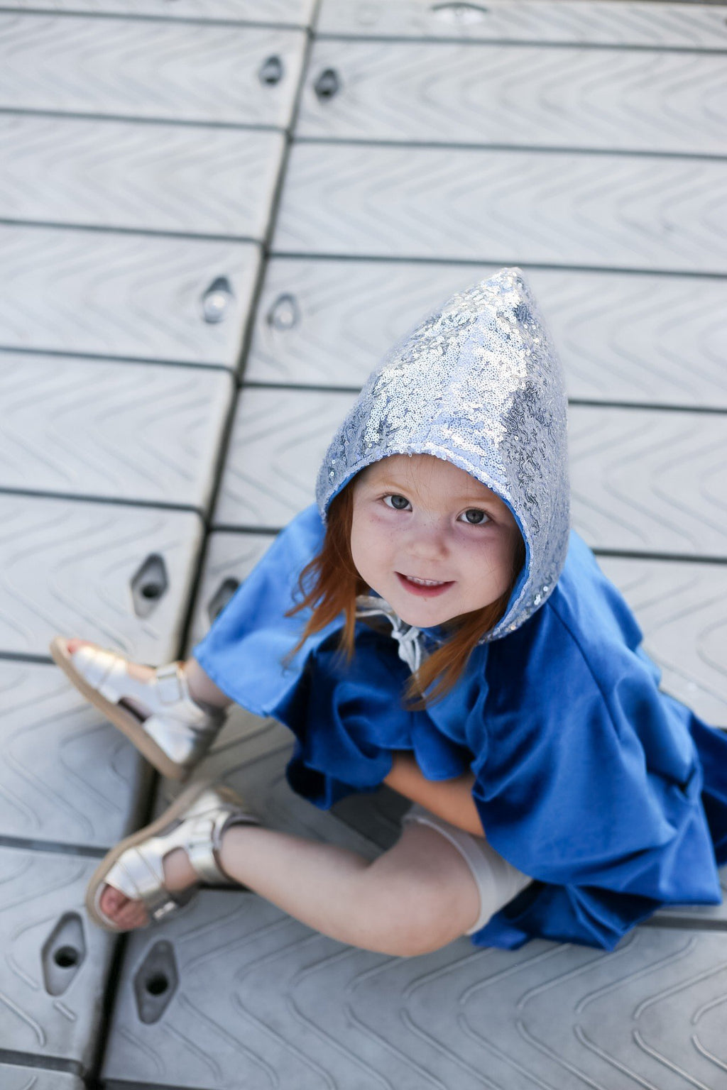5. Child sitting on dock wearing blue cape with silver hood and sandals