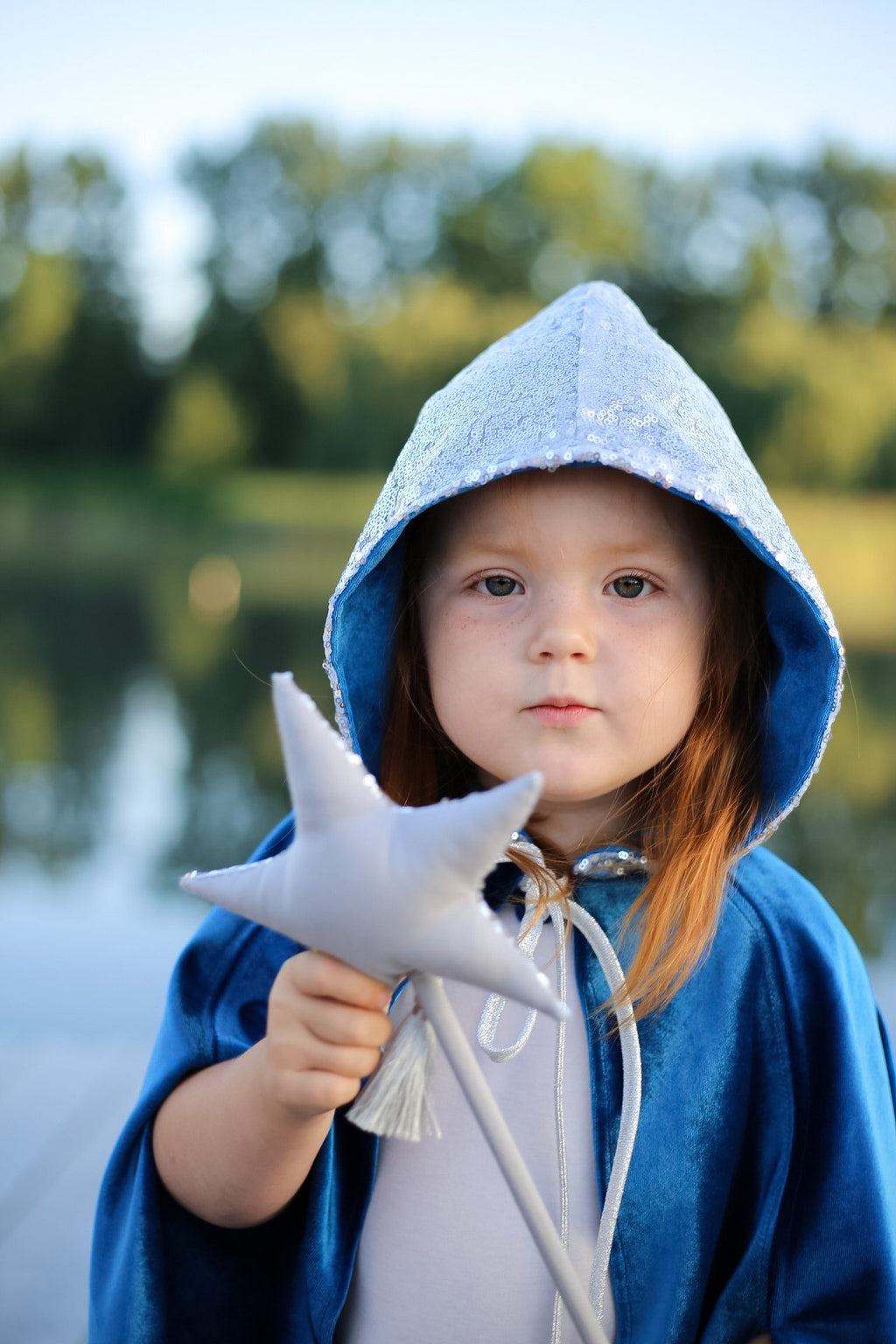 7. Child in blue cape with silver hood holding star wand, standing by a lake