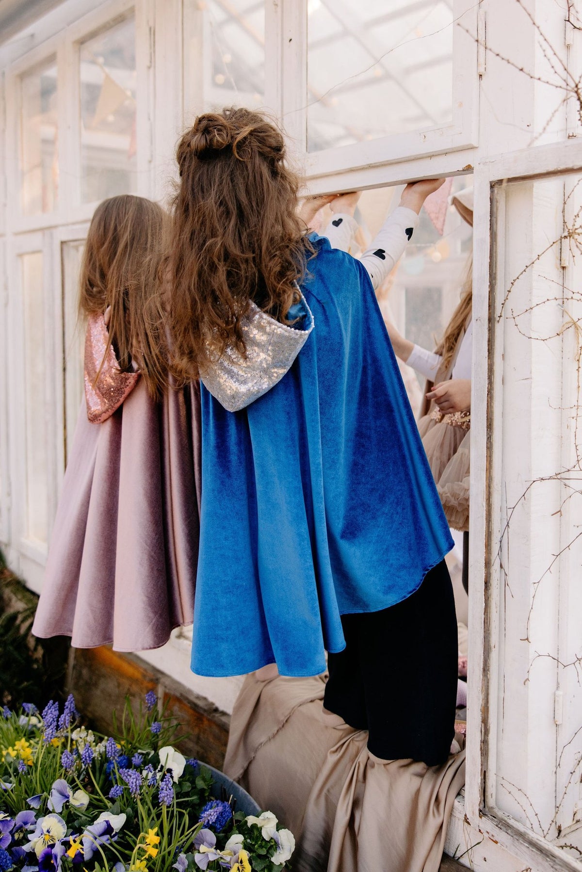 3. Two children wearing blue and pink capes looking out a window in a greenhouse