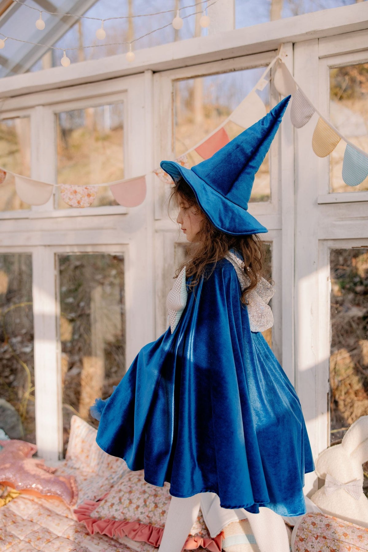 1. Child wearing blue velour cape with pointed hat in a sunlit room, surrounded by colorful cushions and bunting