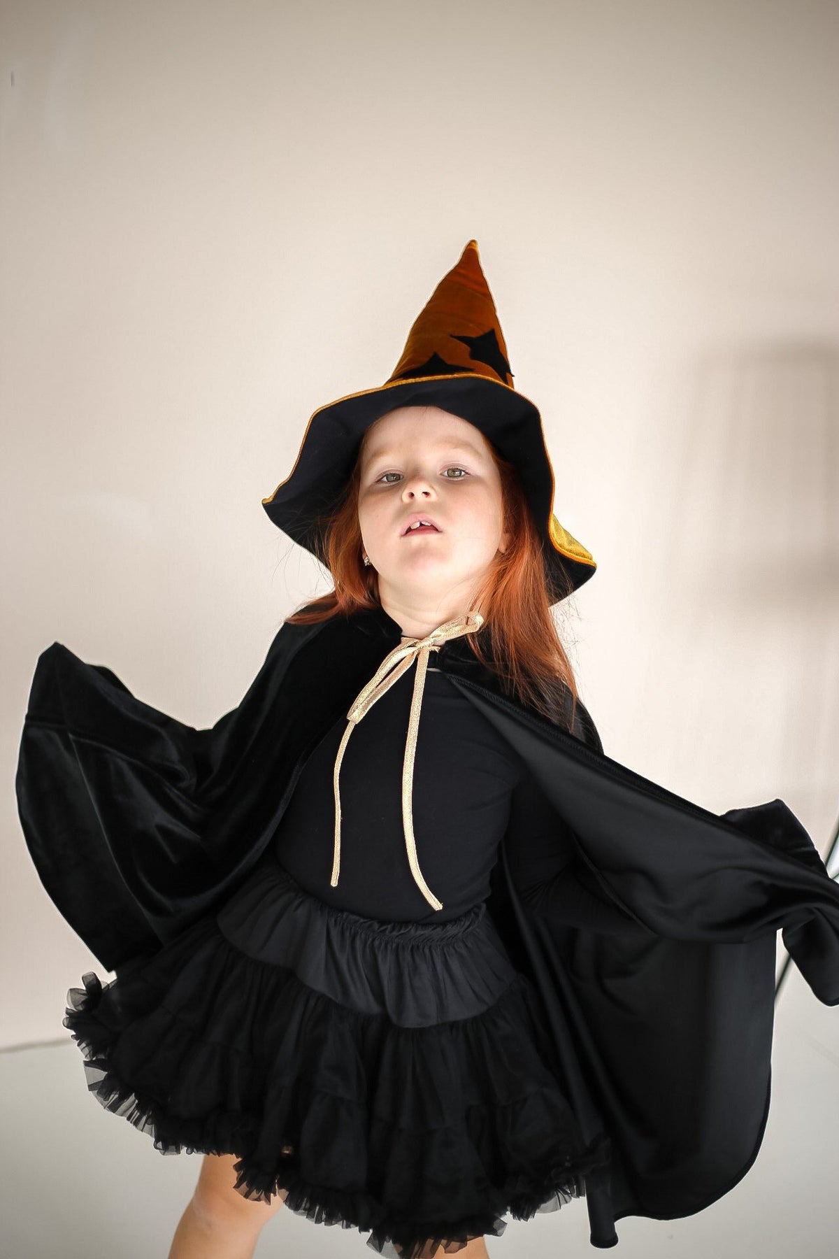 8. Child in black cape and witch hat posing with arms outstretched indoors