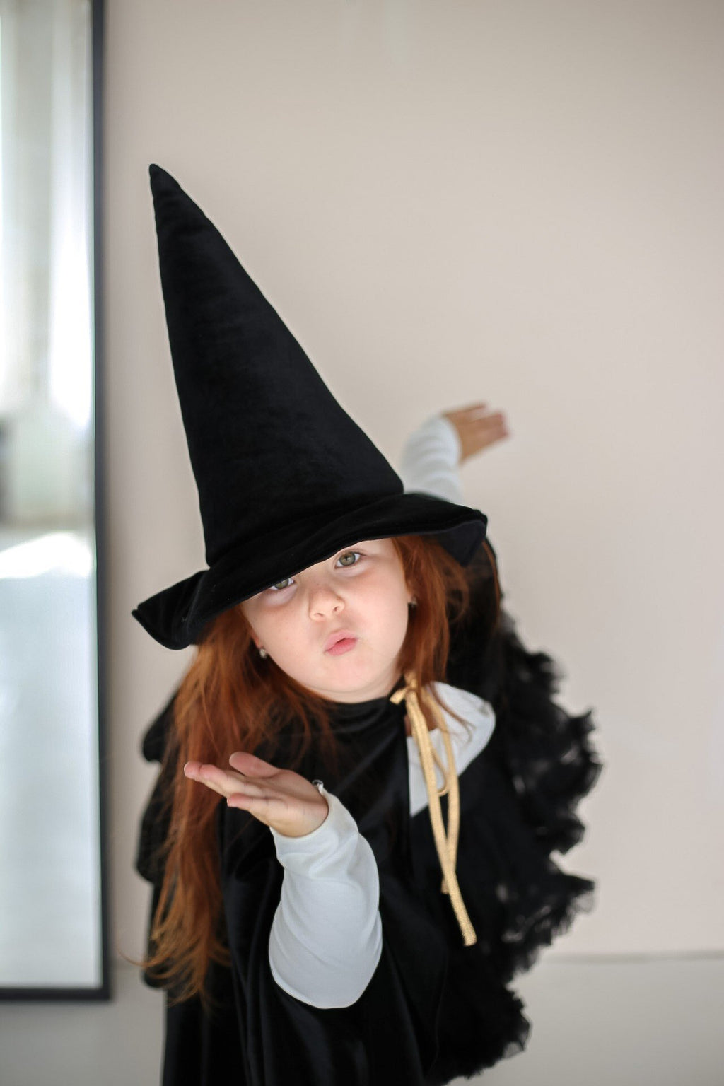 7. Child in black cape and witch hat blowing a kiss indoors