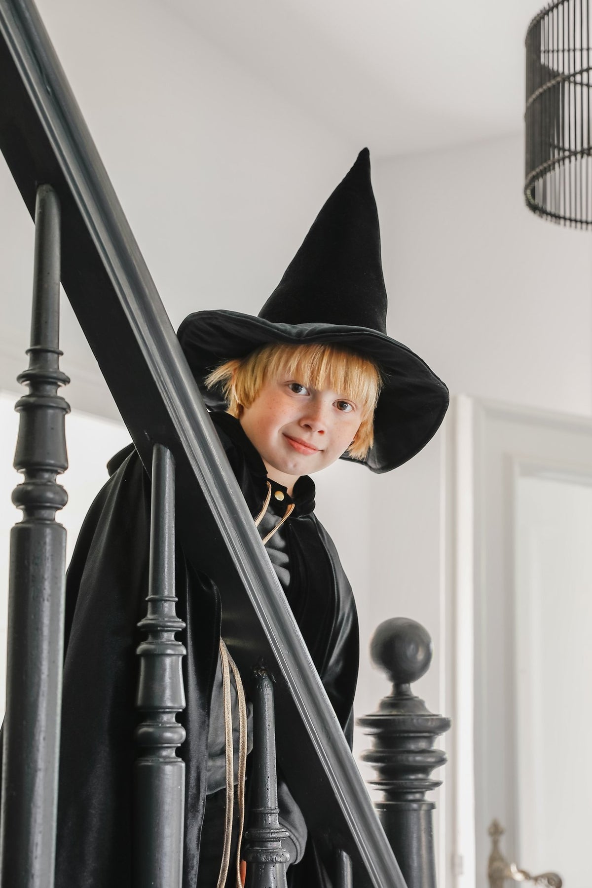 15. Child in black cape and witch hat smiling on staircase indoors