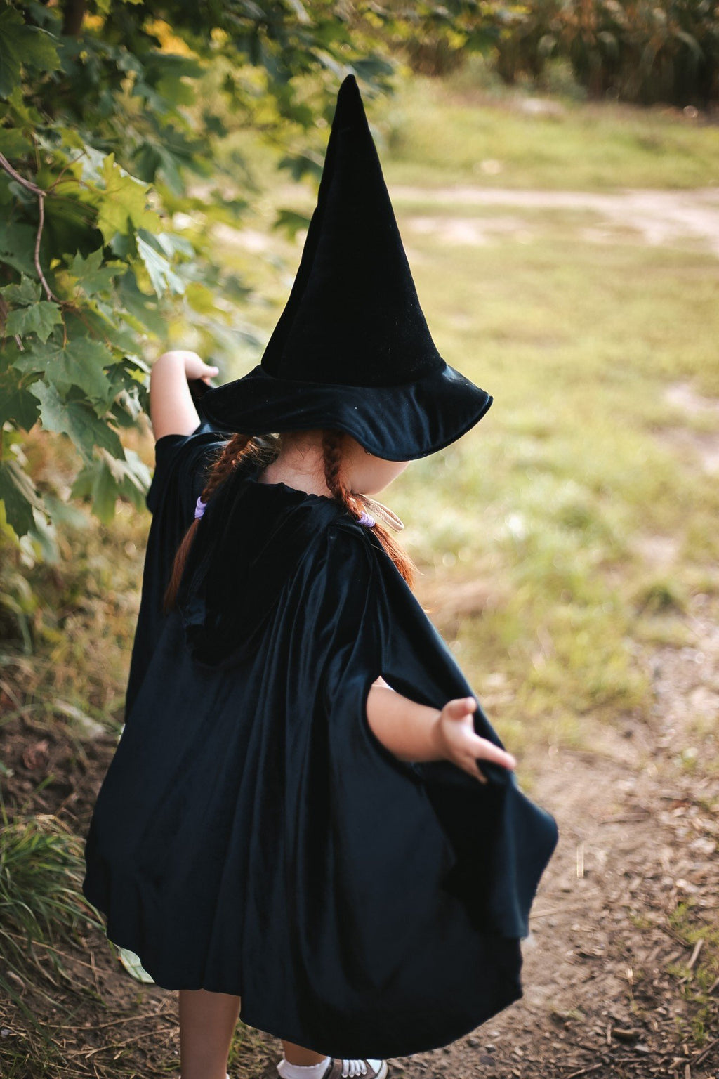 10. Child in black cape and witch hat from back, holding cape in a grassy area