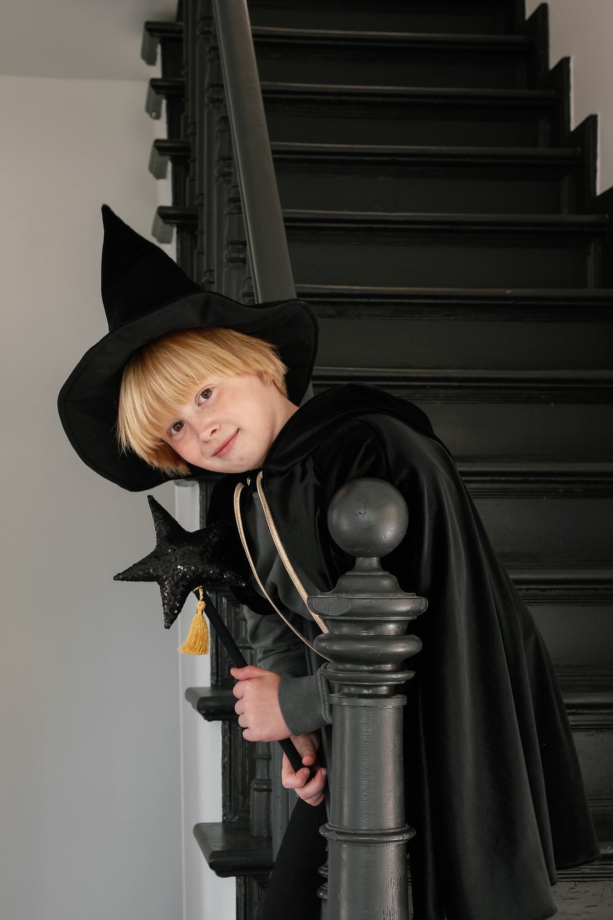 13. Child in black cape and witch hat leaning on staircase railing indoors