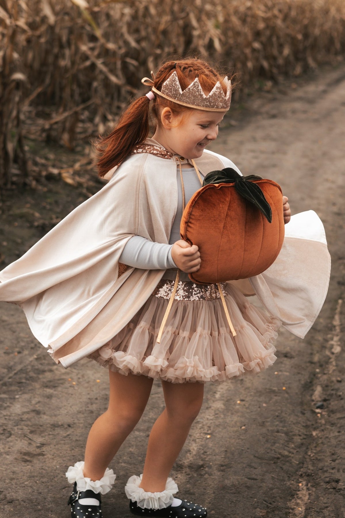 9. Girl in cream cape and crown holding pumpkin on dirt path