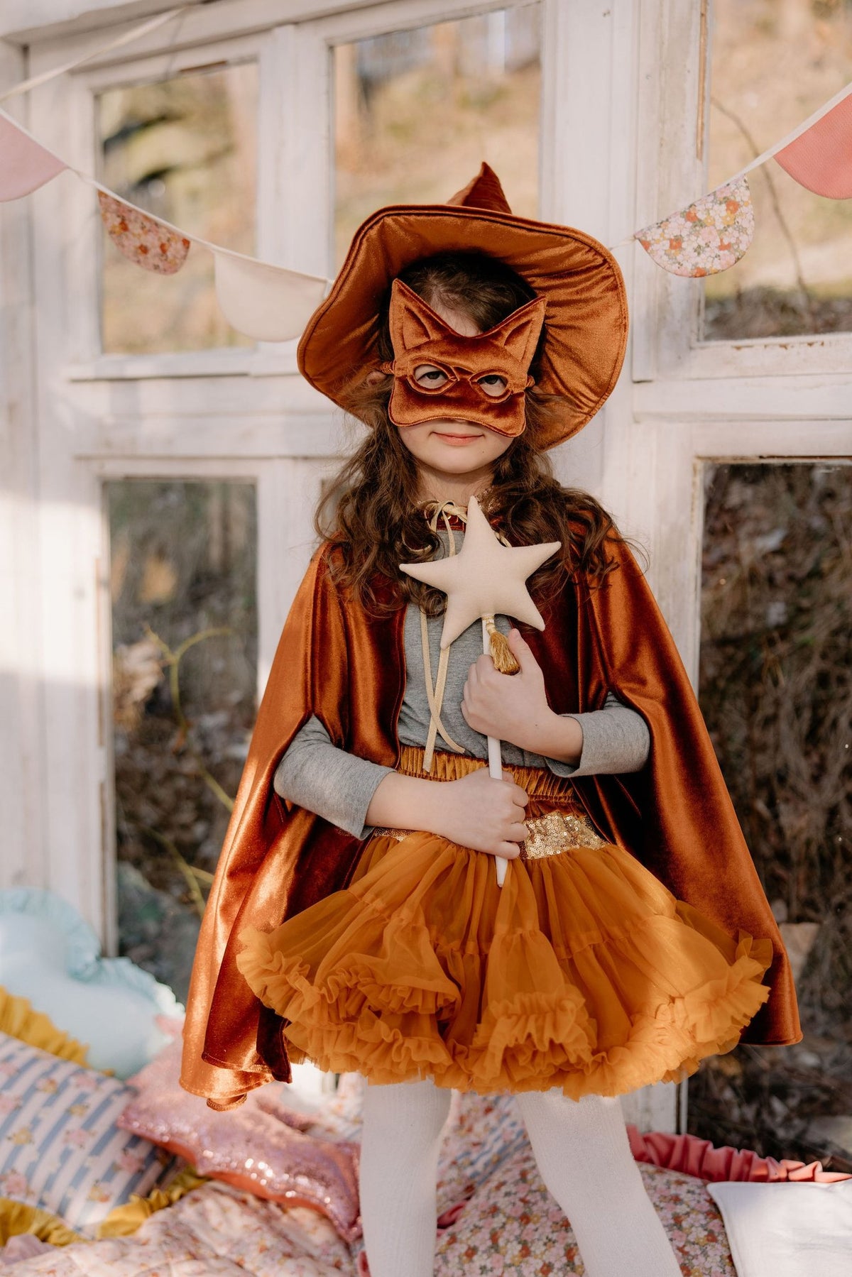 1. Child in ginger cape and mask holding a star wand, standing in a decorated room