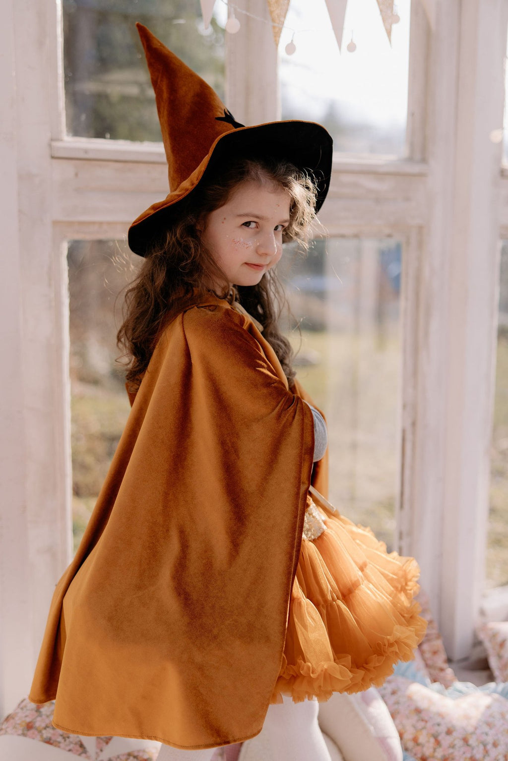 17. Child in golden cape and witch hat sitting by window