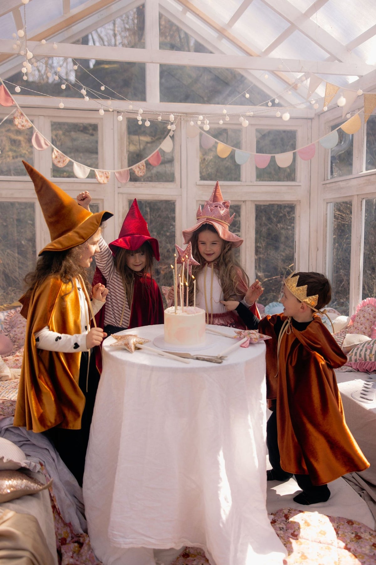 1. Group of children in capes and hats around a table with a cake in a bright room
