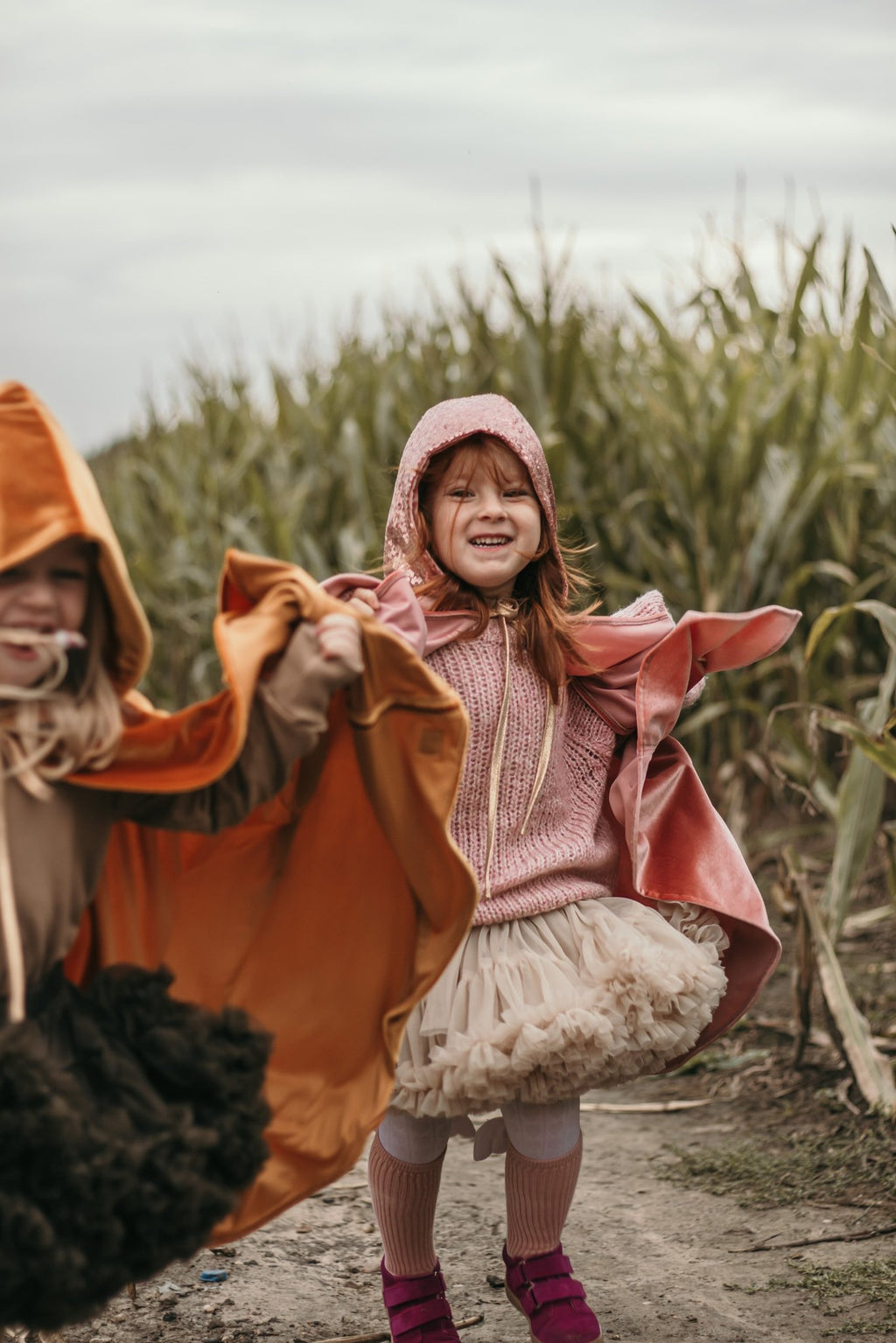 7. Two children in pink and orange capes playing in a cornfield