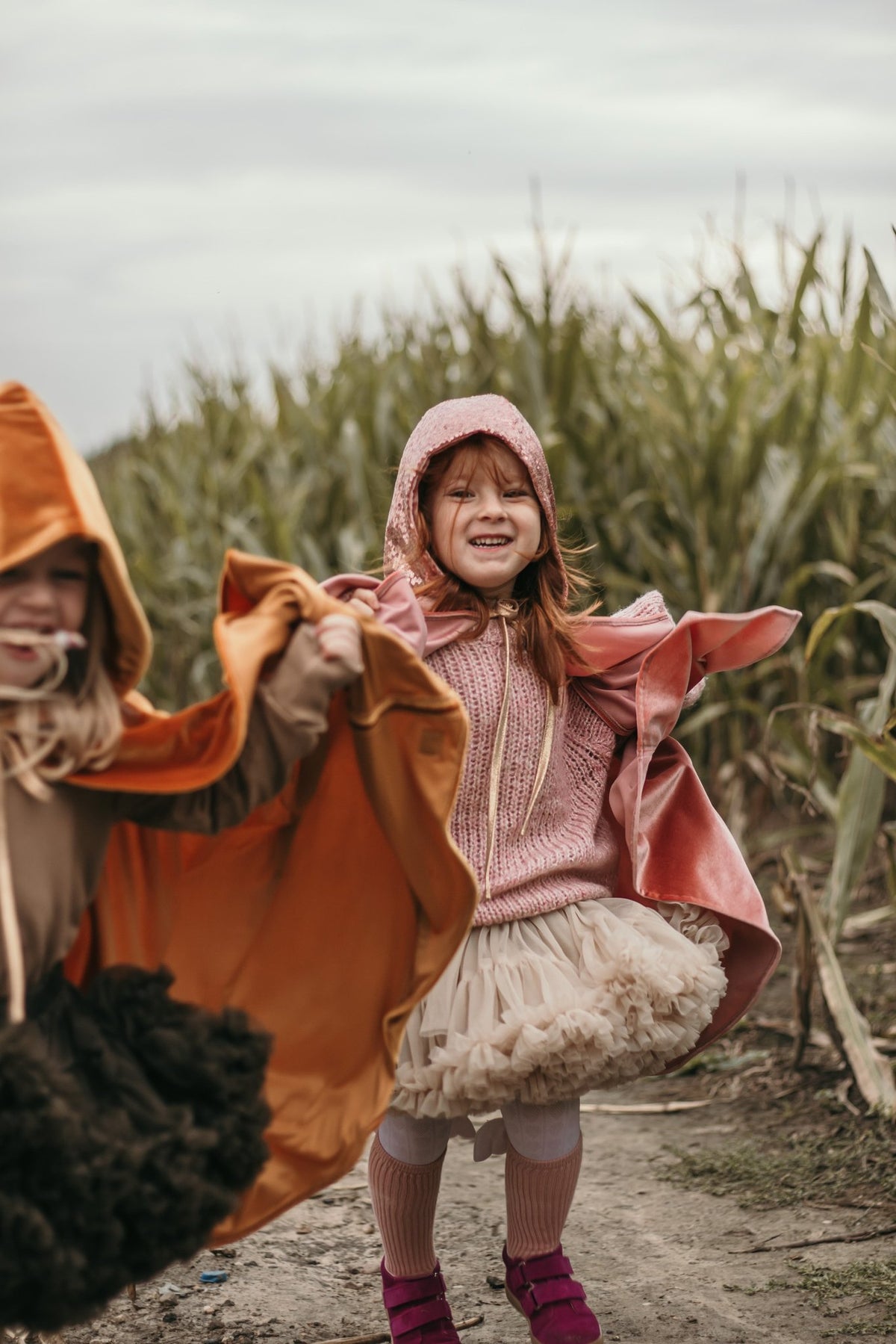 7. Two children in pink and orange capes playing in a cornfield