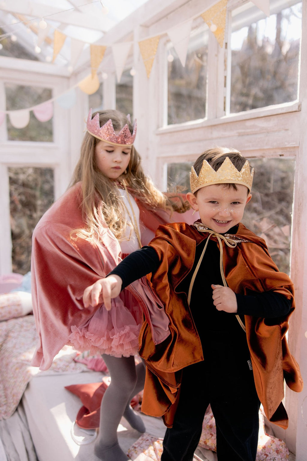 12. Two children in pink and orange capes with crowns playing in a sunlit room