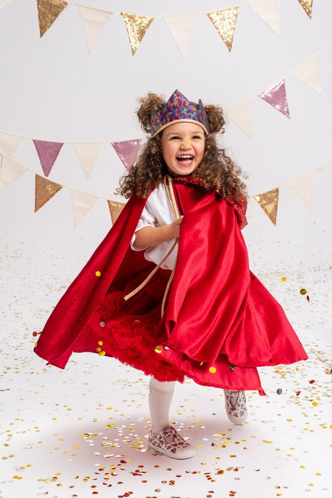 1. Young girl wearing a red velour cape and crown, smiling and playing in a festive room with confetti and bunting decorations