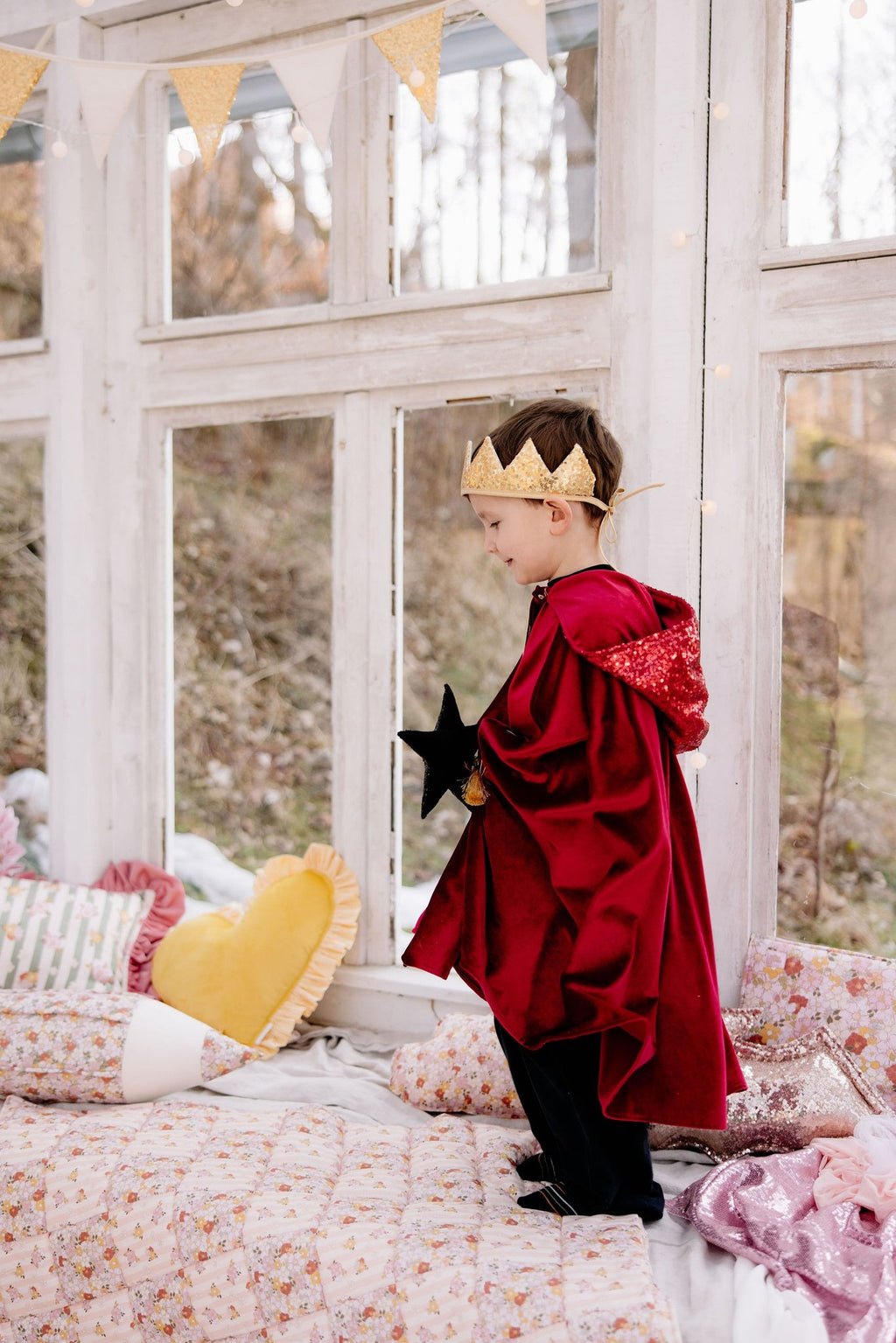 3. Boy wearing a red velour cape and gold crown, standing on a bed holding a star wand, with a scenic outdoor view through large windows